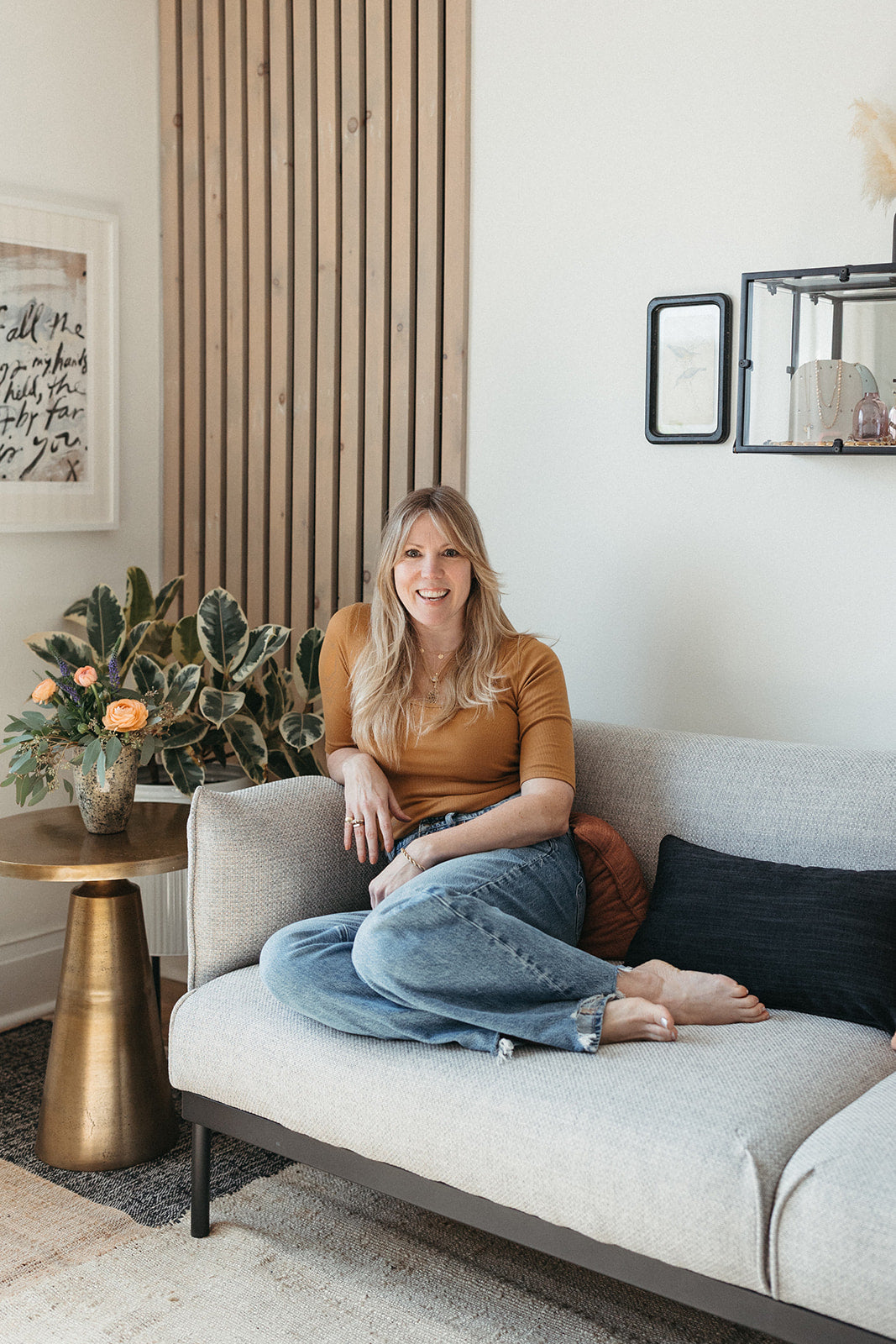 Jilian Maddin sits casually on a beige couch in her Toronto Jewellery studio, smiling warmly. The room has a modern, cozy feel with plants, art, jewellery in glass display cases on the wall and a side table with peachy orange flowers.