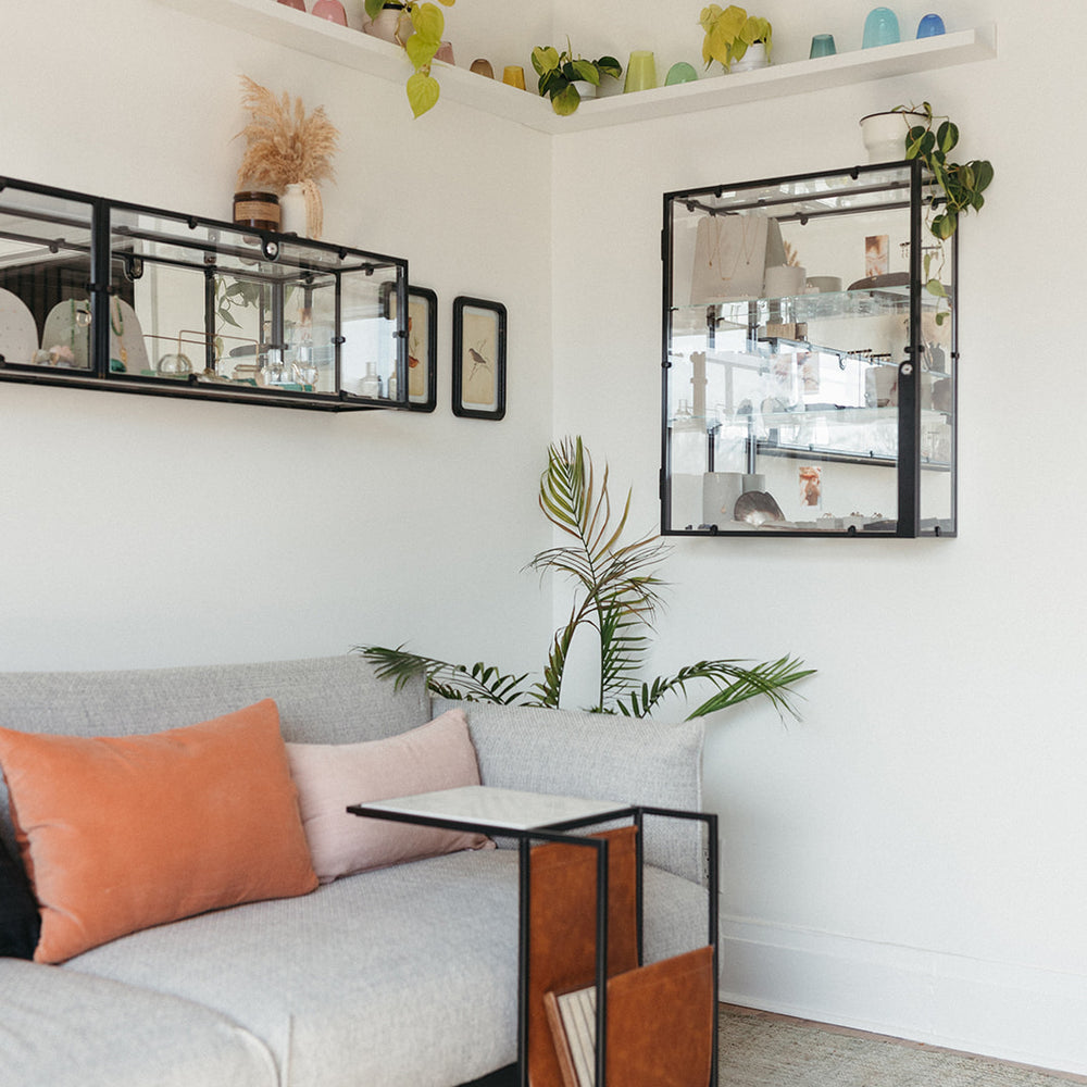 A cozy studio with a gray sofa adorned with orange and pink pillows. A sleek side table holds magazines. Wall shelves display colorful decor and plants.