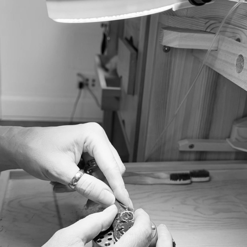 Jilian Maddin's hands wearing rings working on a detailed piece under a magnifying light in her Toronto studio, conveying precision and craftsmanship in a focused setting. The image is in black and white.