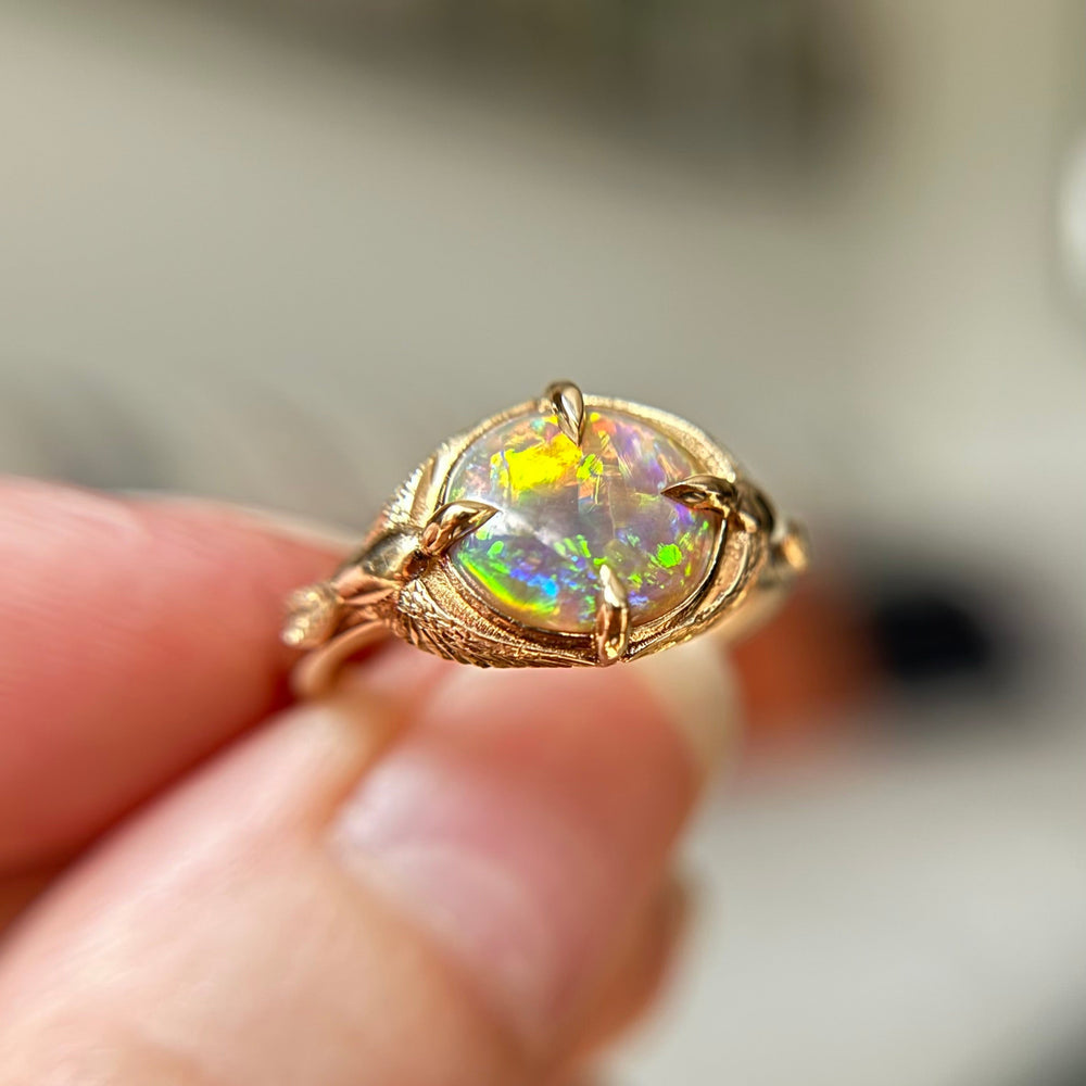 Yellow Gold ring with a beautiful opal centre gemstone, held between fingers against a blurred background. Handmade in Toronto, Canada.
