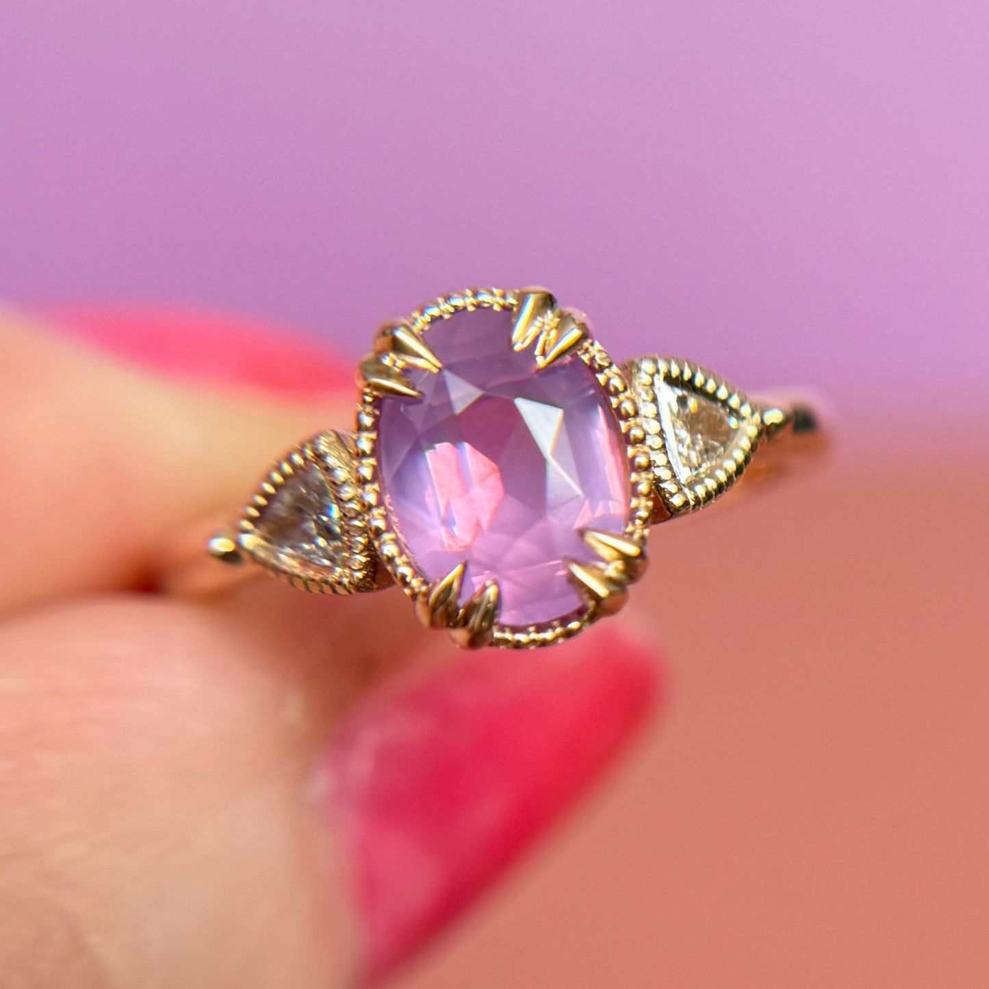 Yellow Gold engagement ring with a luminescent purple sapphire held by a hand against a pink background
