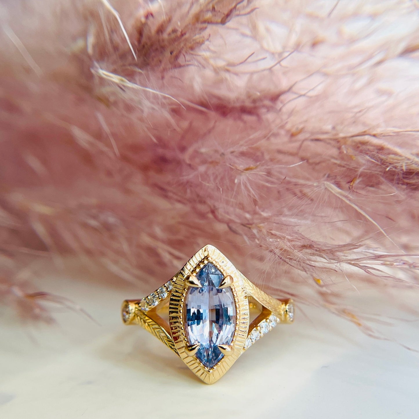 A gold ring with a marquise-shaped pale purple marquise sapphire  and diamonds accents is set against soft, pink pampas grass. The tone is feminine, elegant and luxurious.