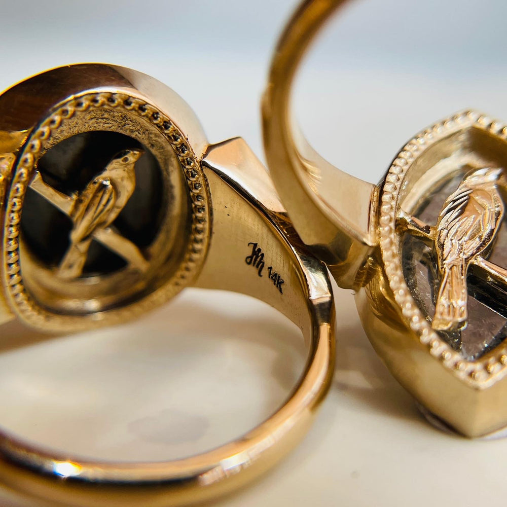 Close-up of two 14K gold rings featuring hand carved bird designs, the bird design is hidden underneath the setting like a unique secret. The engraving "JM 14k" is visible inside the band of one ring.