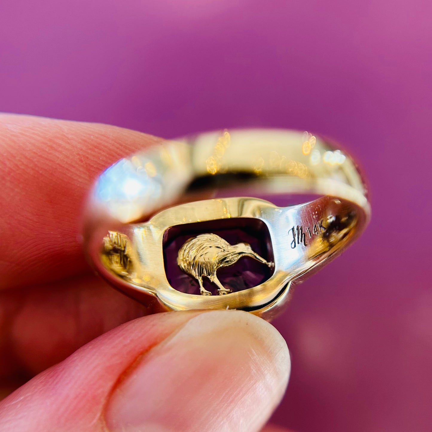 A close-up of a gold ring featuring a hidden engraved kiwi bird under the centre gemstone, held between fingers. Background is a soft purple, giving a warm, elegant feel.