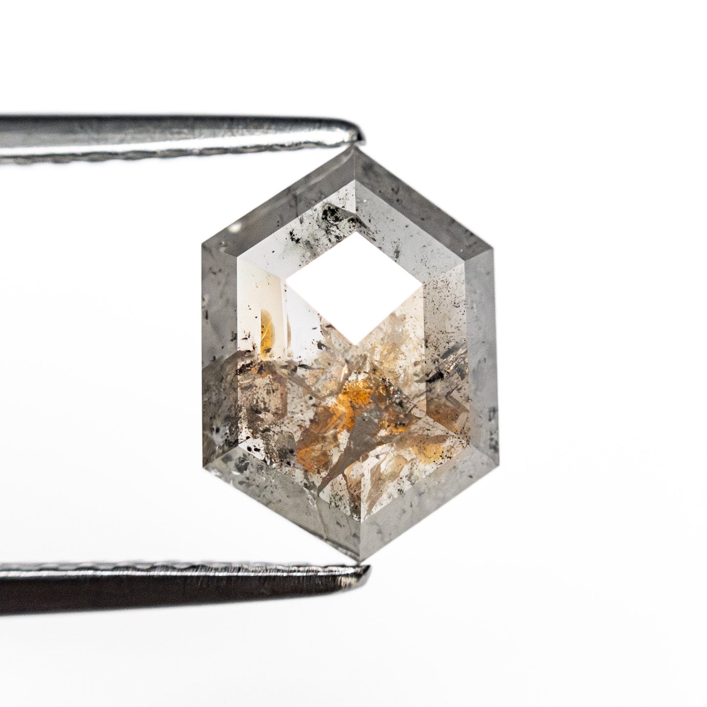 Hexagonal gemstone with a geometric cut, held by metal tweezers. The stone features natural inclusions, with tones of gray and orange, against a white background.