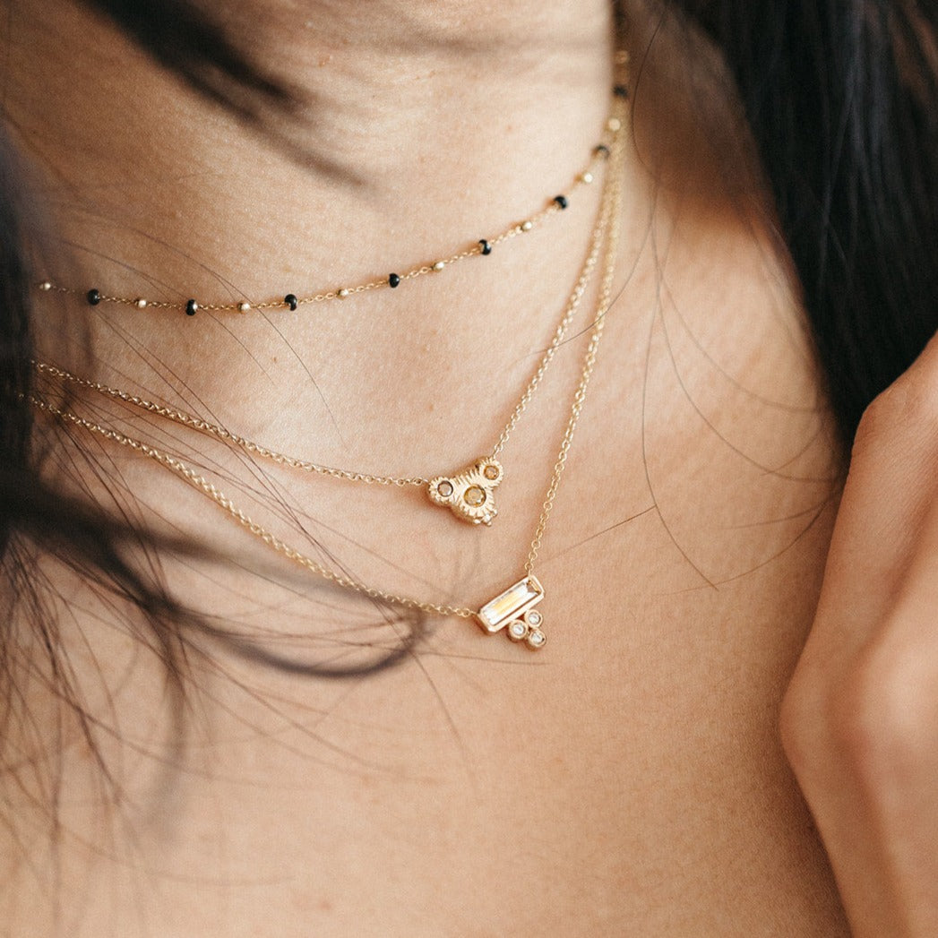 A close-up image of a person wearing a gold necklace with a bar-shaped sunset-colored sapphire and three white diamonds.