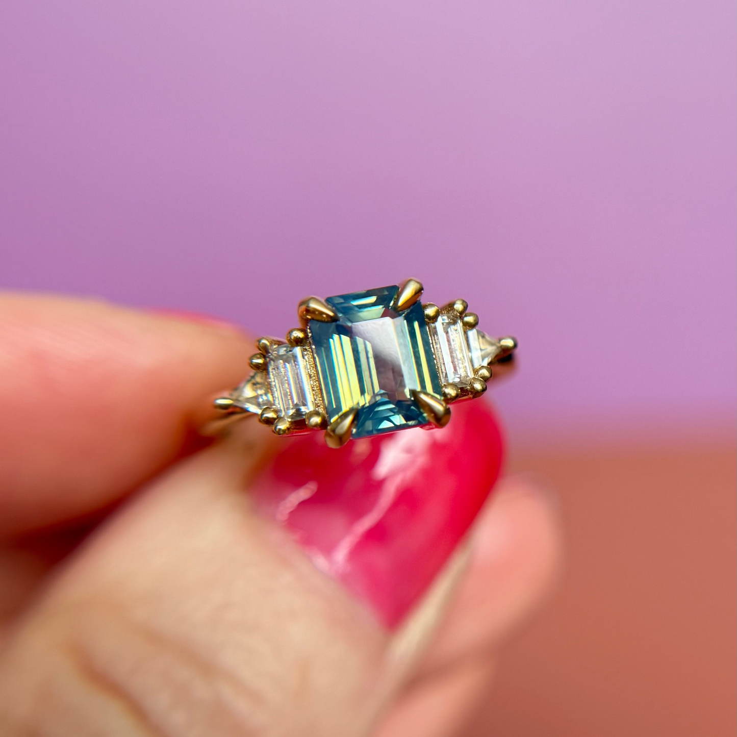 Yellow Gold engagement ring handmade in Toronto Canada with a blue and teal sapphire gemstone held by a hand against a pink background