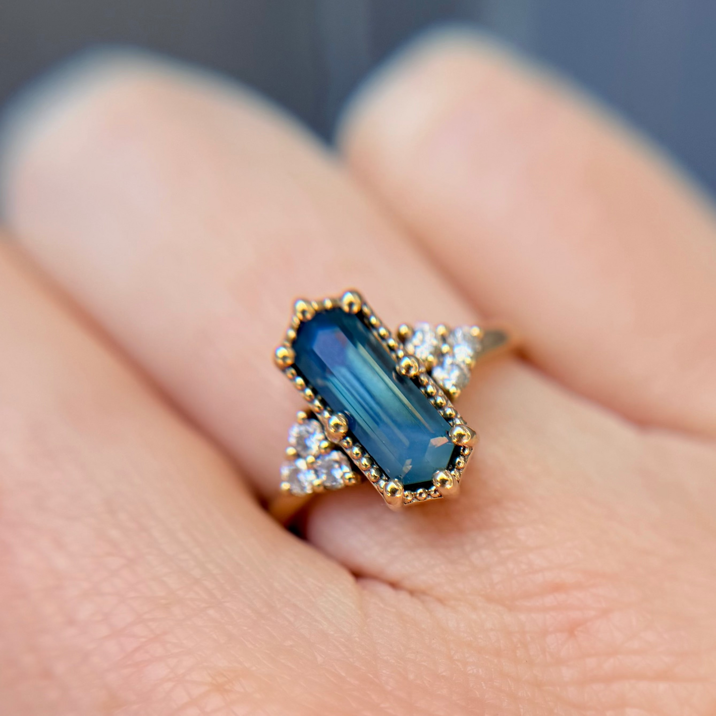 Unique engagement Ring with a blue step cut hexagon sapphire on a finger against a blurred background