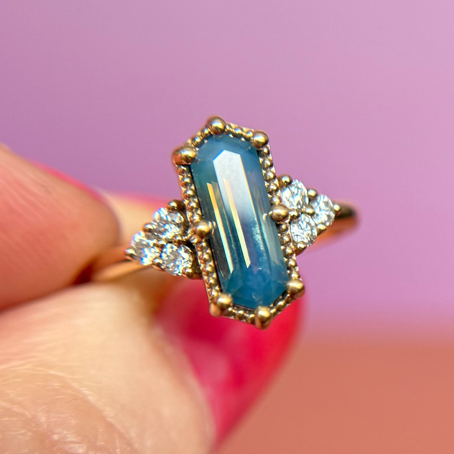 Gold ring with a blue parti or bi-color sapphire and diamond accent stones held between fingers against a blurred background