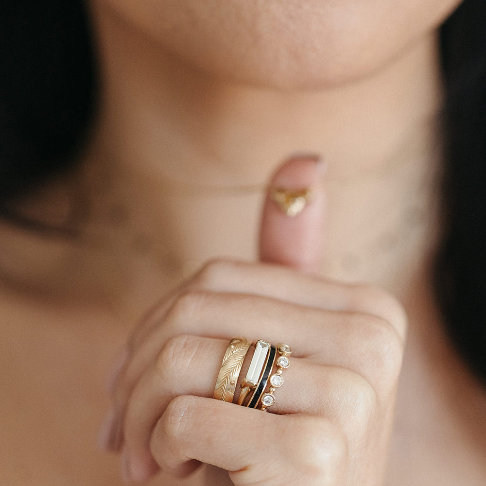 Close-up of a woman's hand touching the necklace around her neck, adorned with gold rings featuring intricate designs and gemstones. The tone is elegant and reflective.