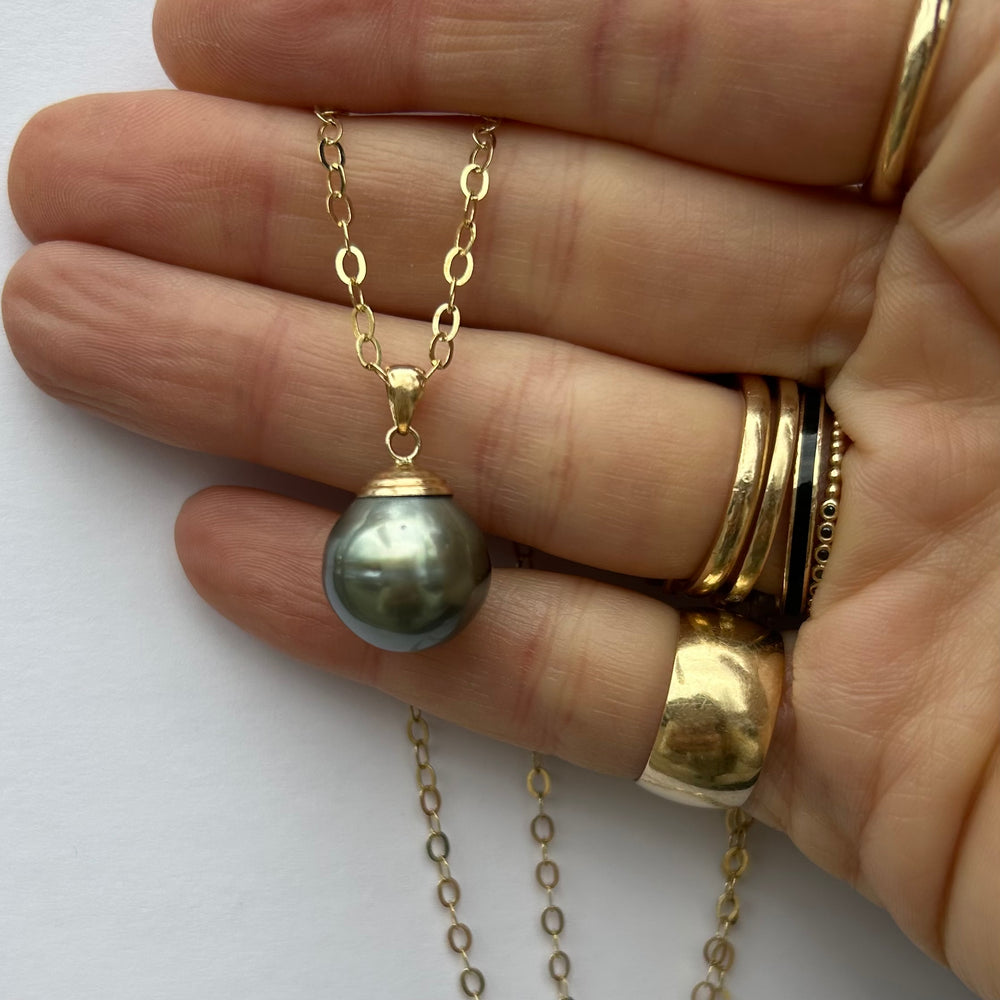 
                  
                    A grey pearl pendant necklace on a 18-inch long yellow gold round cable chain, displayed up close on a hand wearing stacks of yellow gold rings.
                  
                